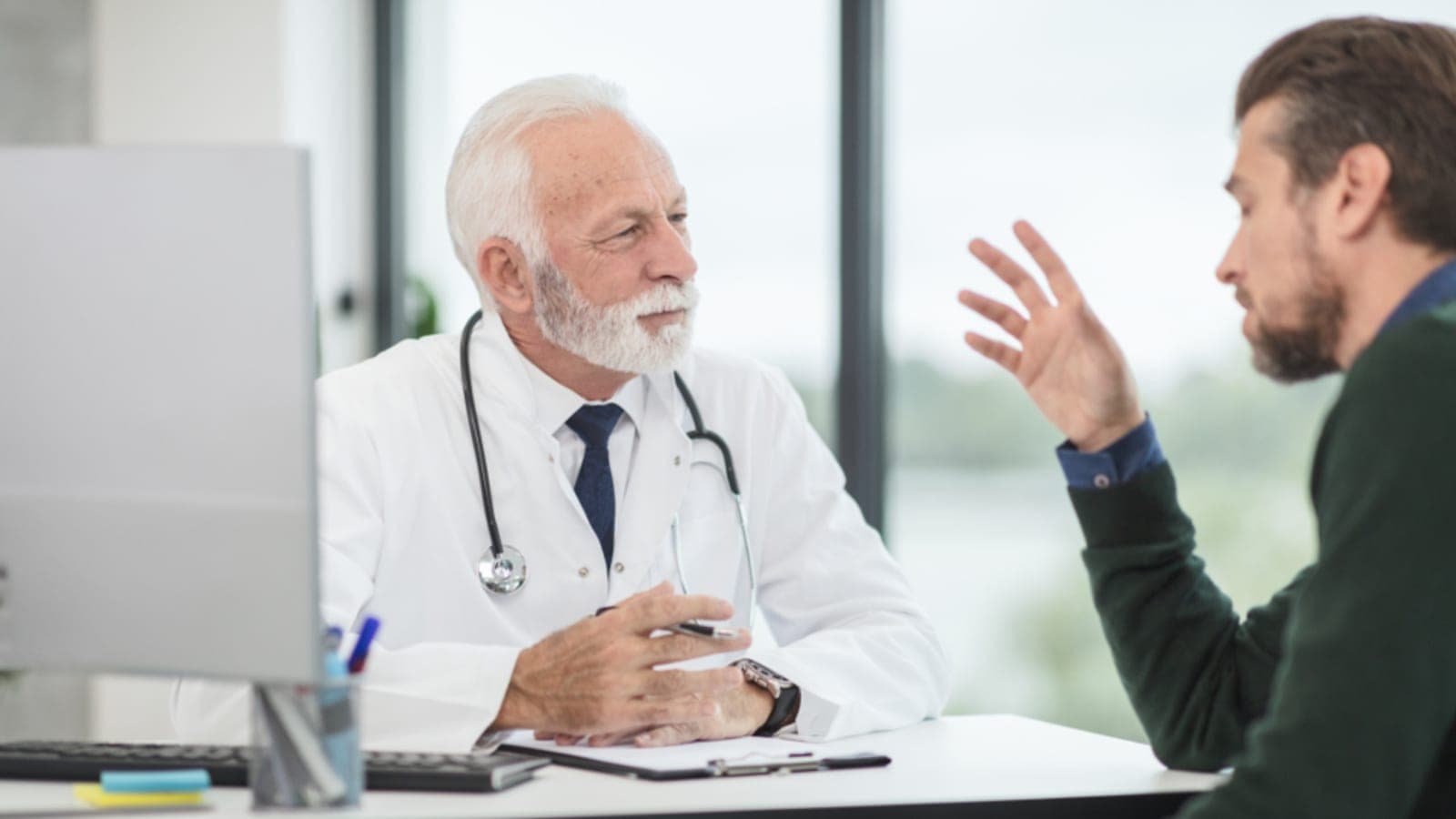 Médico de jaleco branco conversando com um paciente sentado à mesa do consultório